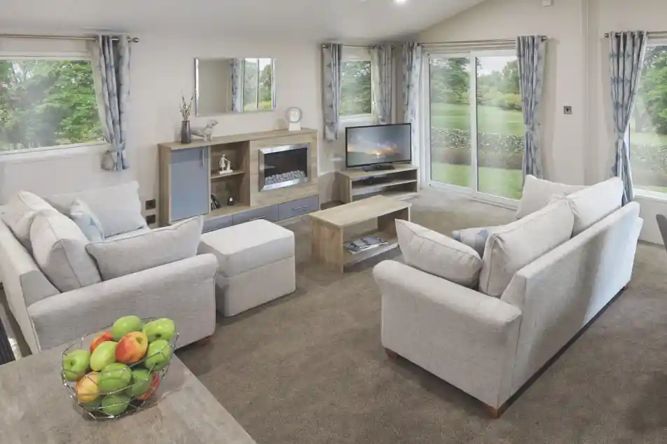 Spacious modern lounge with light grey sofas, a central coffee table, entertainment unit with a TV and electric fireplace, large windows with blue patterned curtains, and sliding glass doors opening to a green garden. A bowl of fresh apples sits on a wooden dining table in the foreground.