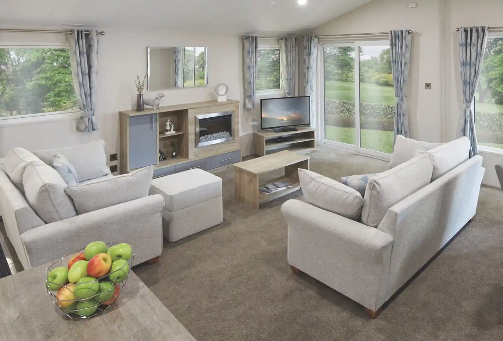 Spacious modern lounge with light grey sofas, a central coffee table, entertainment unit with a TV and electric fireplace, large windows with blue patterned curtains, and sliding glass doors opening to a green garden. A bowl of fresh apples sits on a wooden dining table in the foreground.