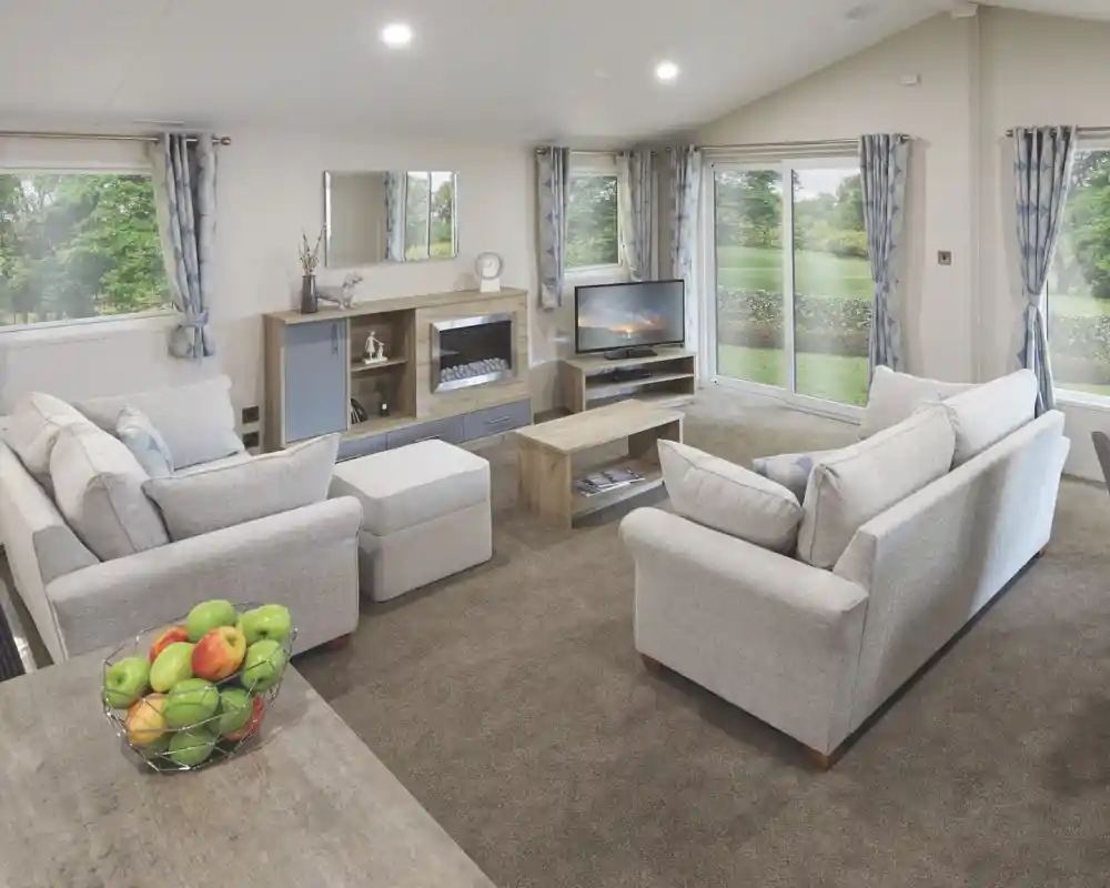Spacious modern lounge with light grey sofas, a central coffee table, entertainment unit with a TV and electric fireplace, large windows with blue patterned curtains, and sliding glass doors opening to a green garden. A bowl of fresh apples sits on a wooden dining table in the foreground.