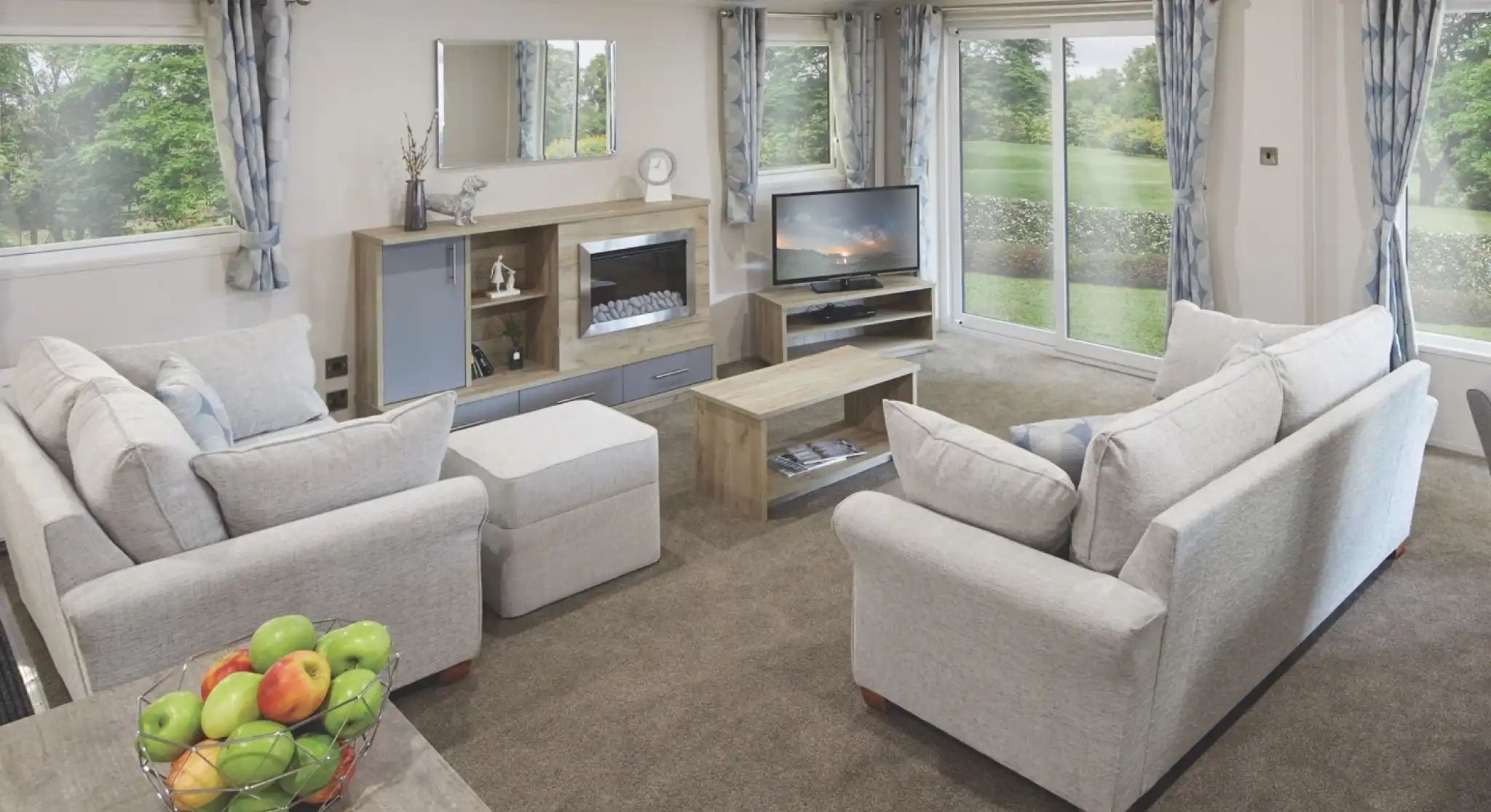 Spacious modern lounge with light grey sofas, a central coffee table, entertainment unit with a TV and electric fireplace, large windows with blue patterned curtains, and sliding glass doors opening to a green garden. A bowl of fresh apples sits on a wooden dining table in the foreground.