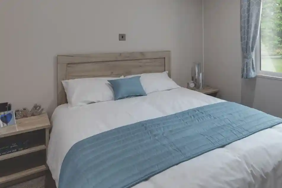 Cozy master bedroom with a wooden headboard, white bedding, and a light blue throw and pillow. Two bedside tables hold decorative items, a clock, and books. A window with patterned blue curtains lets in natural light and offers a view of the green outdoors.