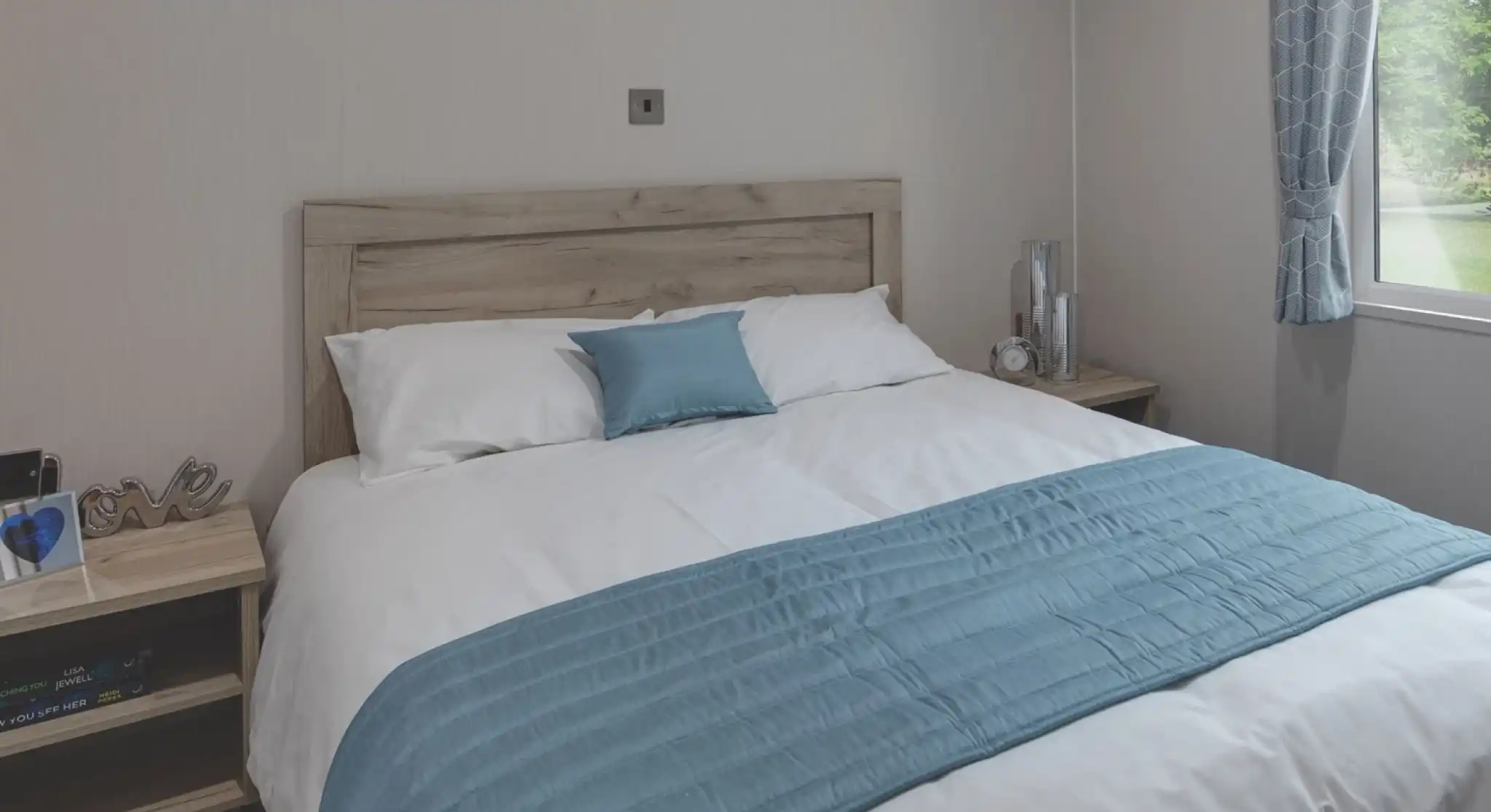 Cozy master bedroom with a wooden headboard, white bedding, and a light blue throw and pillow. Two bedside tables hold decorative items, a clock, and books. A window with patterned blue curtains lets in natural light and offers a view of the green outdoors.