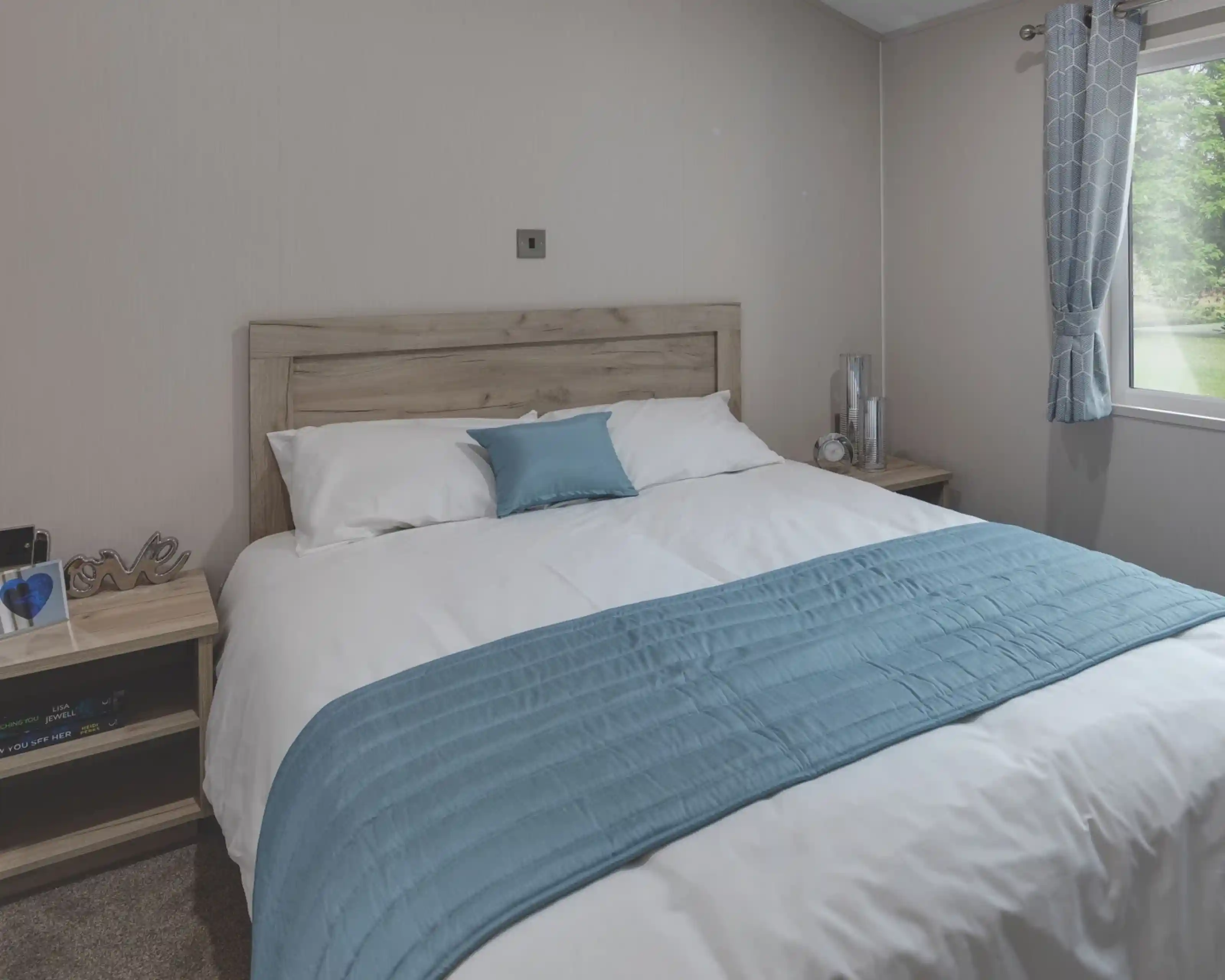 Cozy master bedroom with a wooden headboard, white bedding, and a light blue throw and pillow. Two bedside tables hold decorative items, a clock, and books. A window with patterned blue curtains lets in natural light and offers a view of the green outdoors.