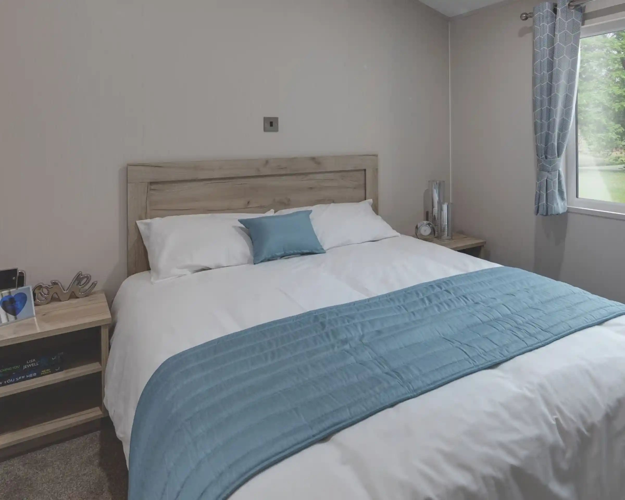 Cozy master bedroom with a wooden headboard, white bedding, and a light blue throw and pillow. Two bedside tables hold decorative items, a clock, and books. A window with patterned blue curtains lets in natural light and offers a view of the green outdoors.