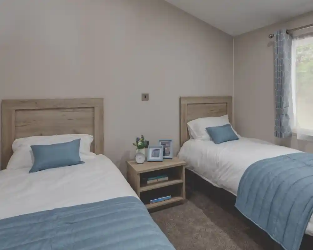 Twin bedroom with two single beds separated by a wooden nightstand. Each bed has white bedding and a light blue throw blanket and pillow. The nightstand holds two photo frames, a small clock, and a vase with flowers. A window with blue patterned curtains allows natural light into the room.