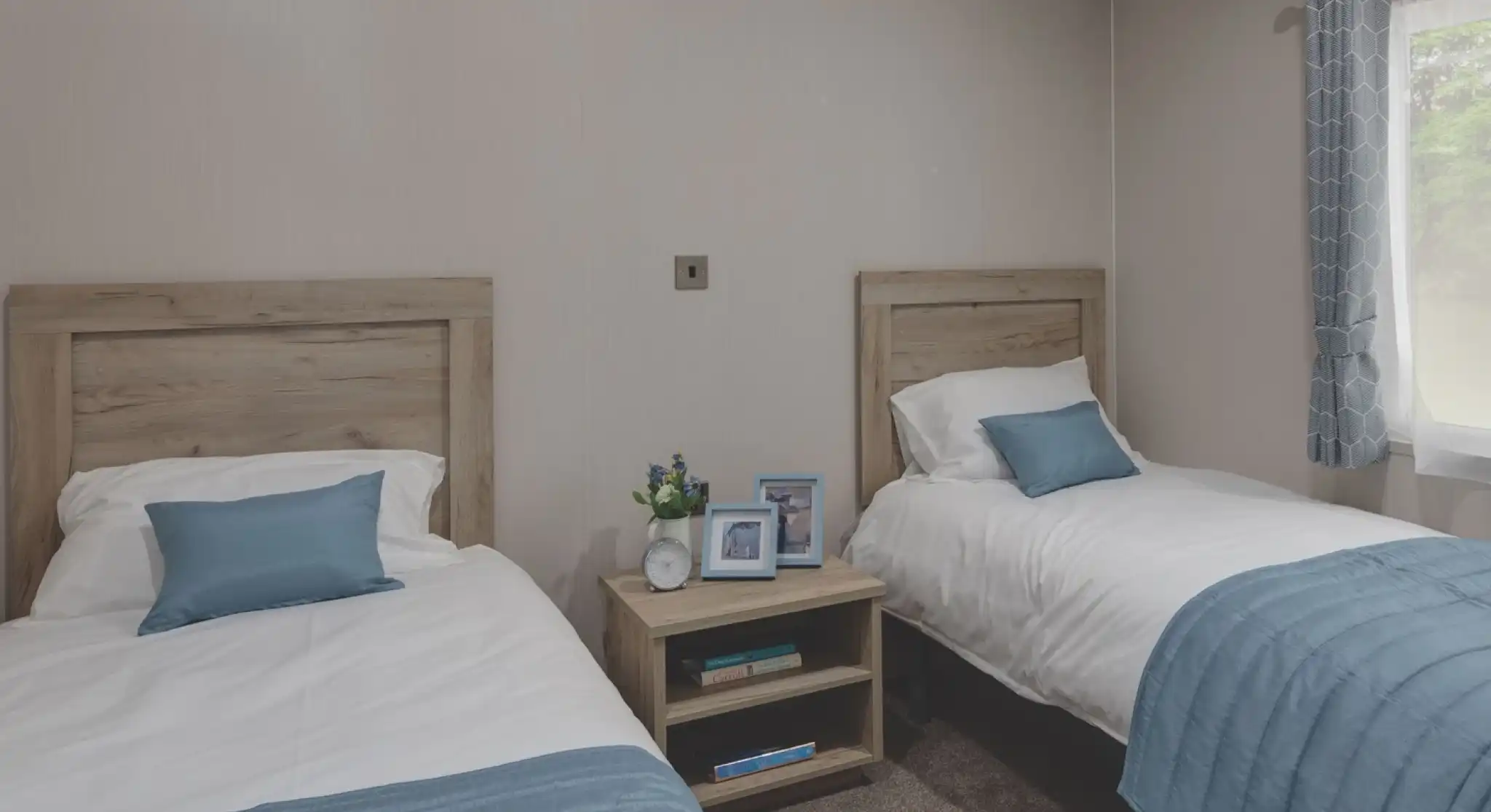 Twin bedroom with two single beds separated by a wooden nightstand. Each bed has white bedding and a light blue throw blanket and pillow. The nightstand holds two photo frames, a small clock, and a vase with flowers. A window with blue patterned curtains allows natural light into the room.