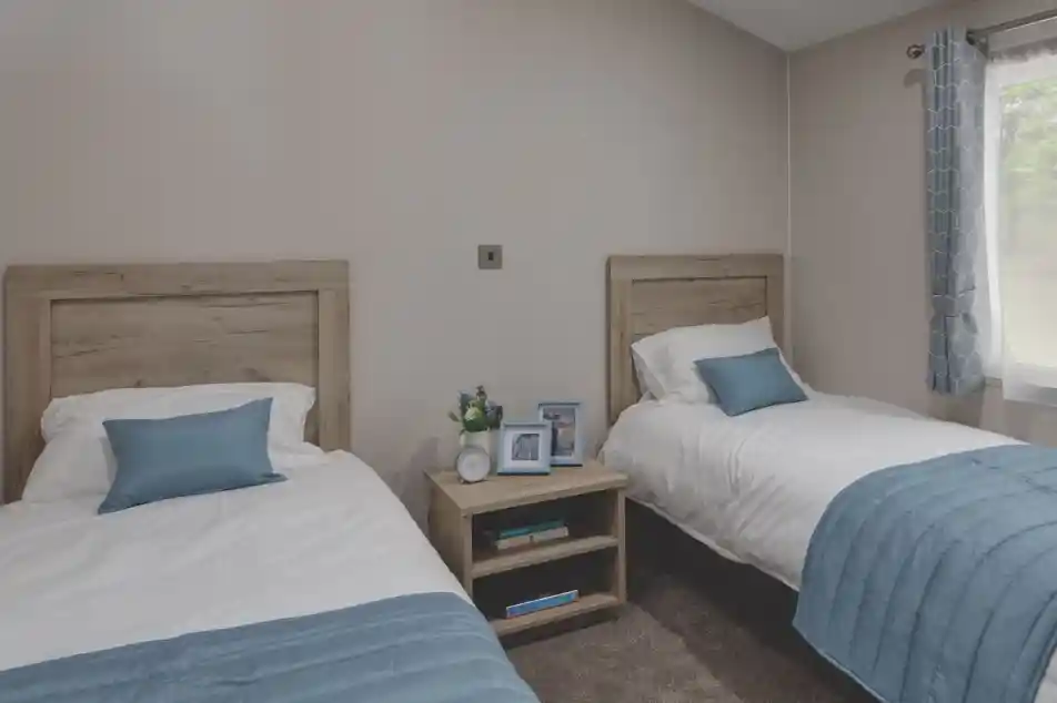 Twin bedroom with two single beds separated by a wooden nightstand. Each bed has white bedding and a light blue throw blanket and pillow. The nightstand holds two photo frames, a small clock, and a vase with flowers. A window with blue patterned curtains allows natural light into the room.