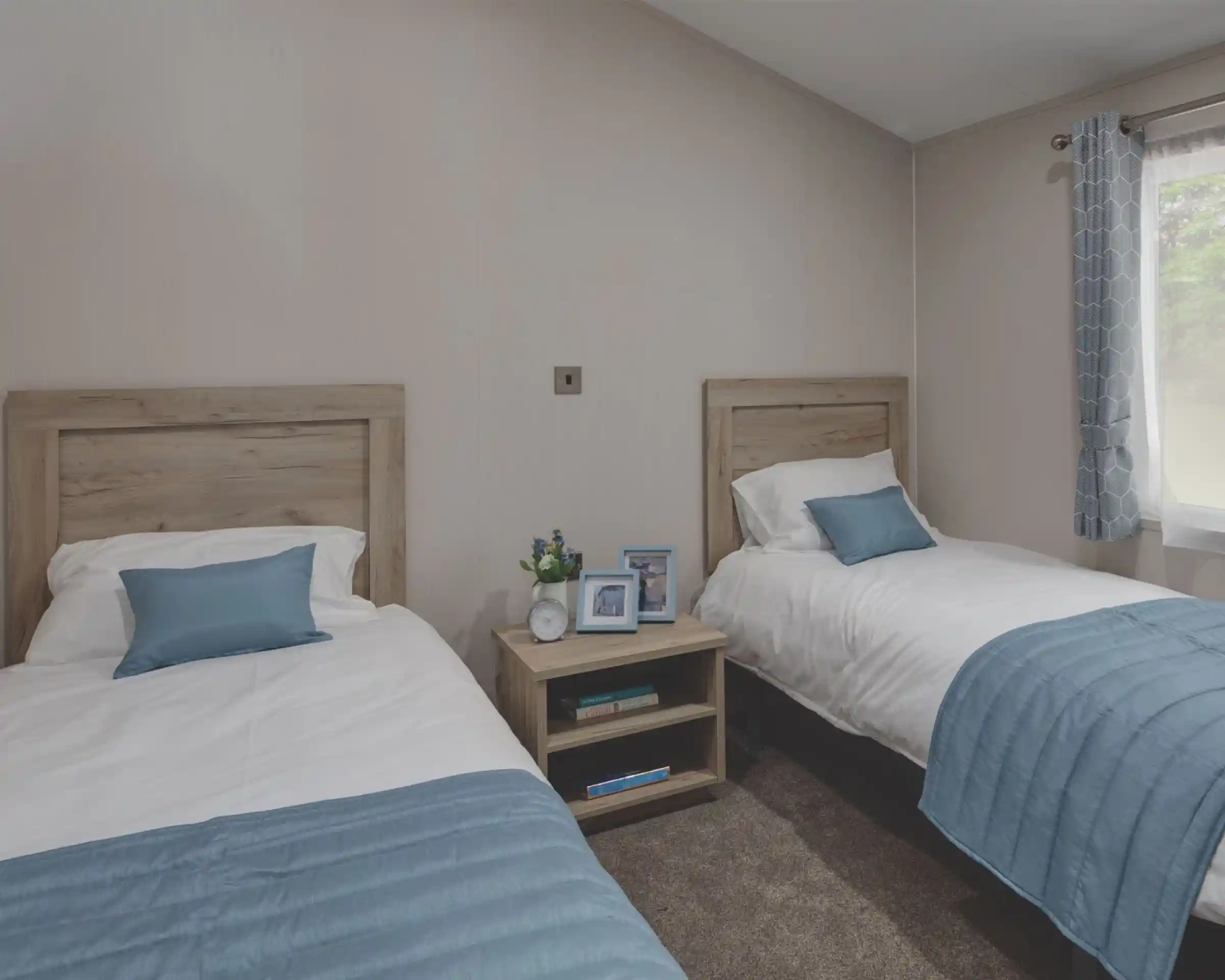 Twin bedroom with two single beds separated by a wooden nightstand. Each bed has white bedding and a light blue throw blanket and pillow. The nightstand holds two photo frames, a small clock, and a vase with flowers. A window with blue patterned curtains allows natural light into the room.