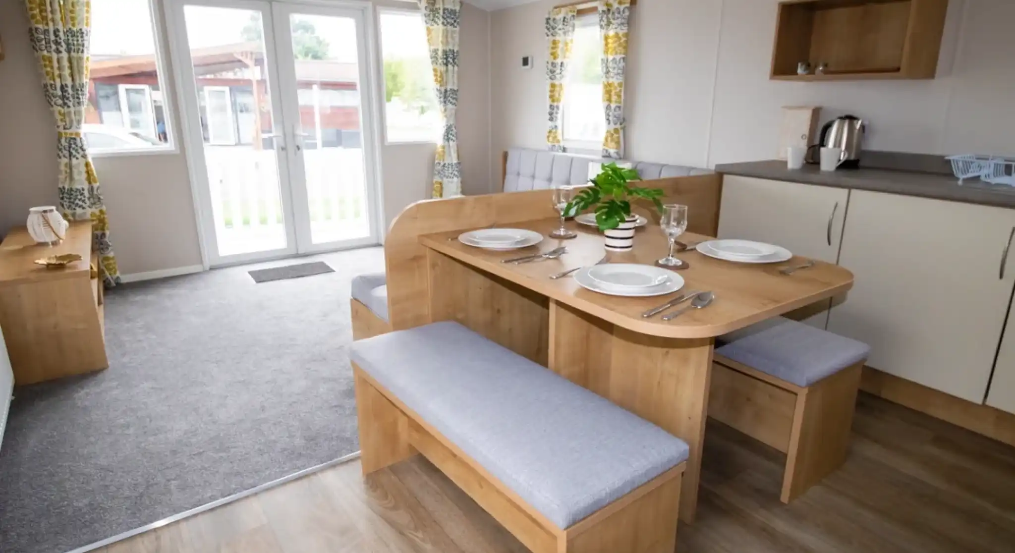 A cozy dining area featuring a wooden table with gray upholstered benches set for a meal. Large windows with patterned curtains allow natural light, and a small plant sits center table. A kitchenette is visible in the background with a kettle and cabinets.