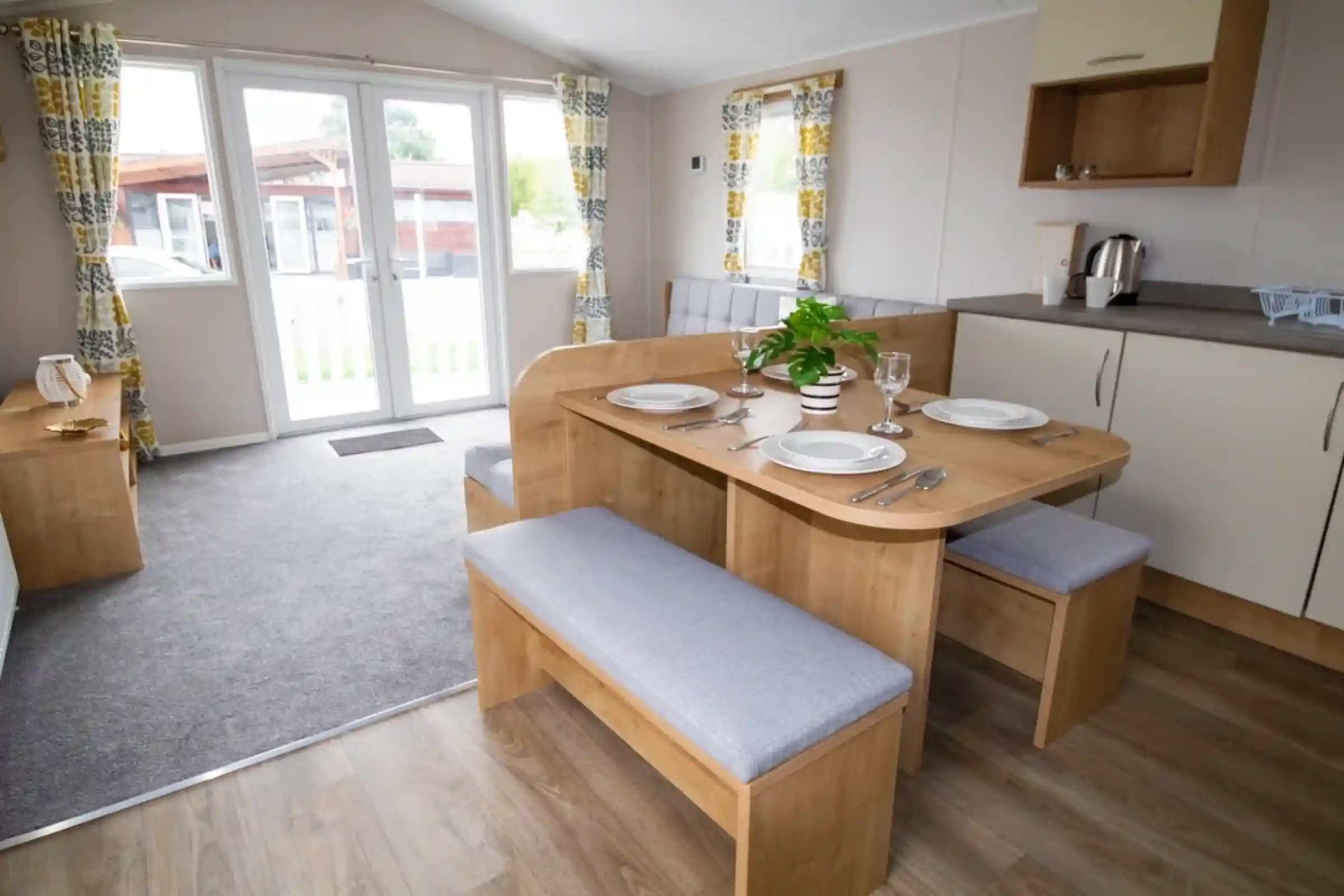A cozy dining area featuring a wooden table with gray upholstered benches set for a meal. Large windows with patterned curtains allow natural light, and a small plant sits center table. A kitchenette is visible in the background with a kettle and cabinets.