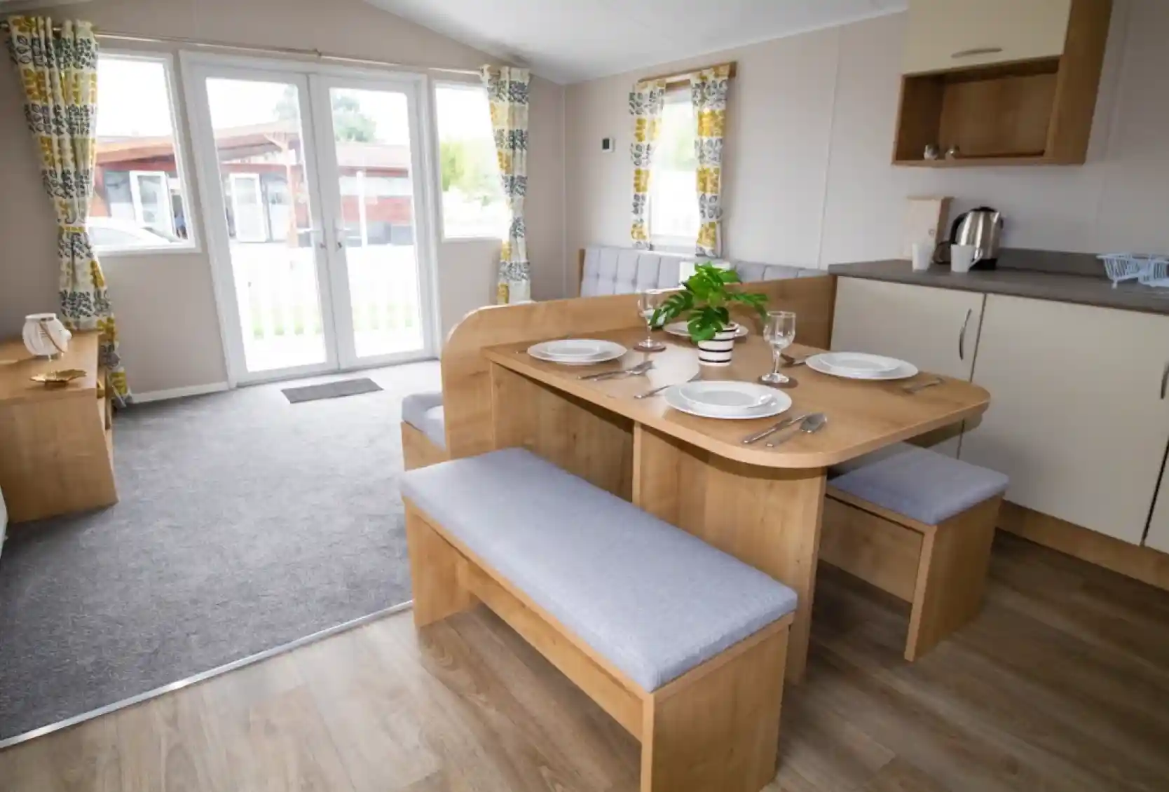 A cozy dining area featuring a wooden table with gray upholstered benches set for a meal. Large windows with patterned curtains allow natural light, and a small plant sits center table. A kitchenette is visible in the background with a kettle and cabinets.