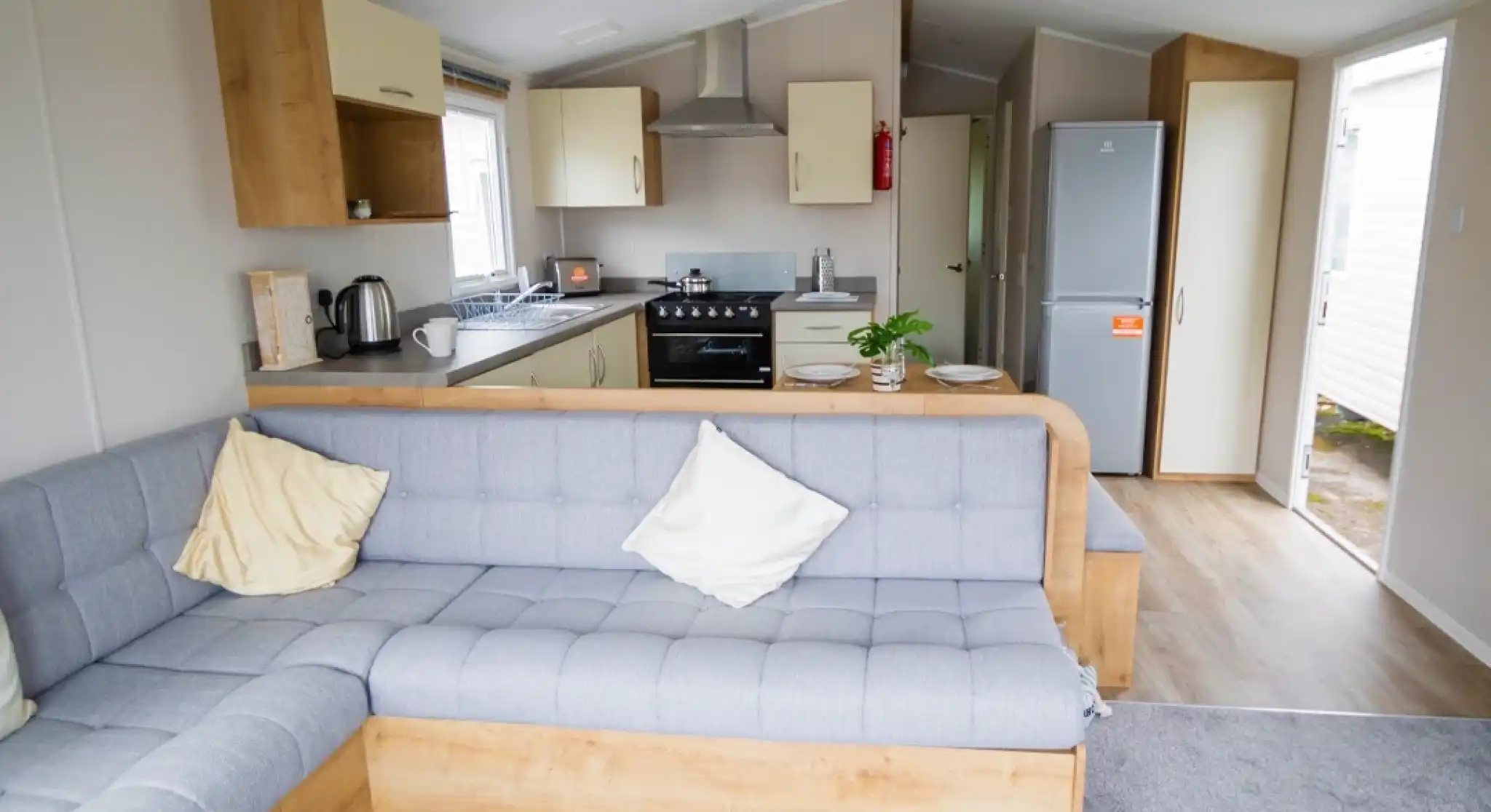 Modern interior of a cozy mobile home featuring a gray sectional sofa with decorative pillows, a compact kitchen with a gas stove and refrigerator, and light wood cabinetry. Natural light enters through the windows, creating a welcoming atmosphere.