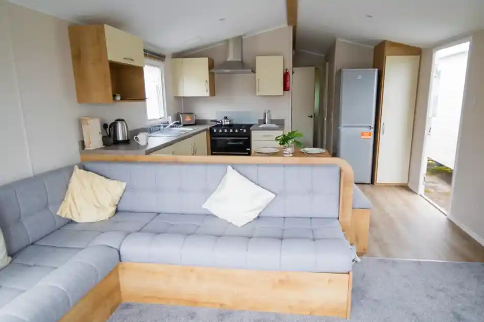 Modern interior of a cozy mobile home featuring a gray sectional sofa with decorative pillows, a compact kitchen with a gas stove and refrigerator, and light wood cabinetry. Natural light enters through the windows, creating a welcoming atmosphere.