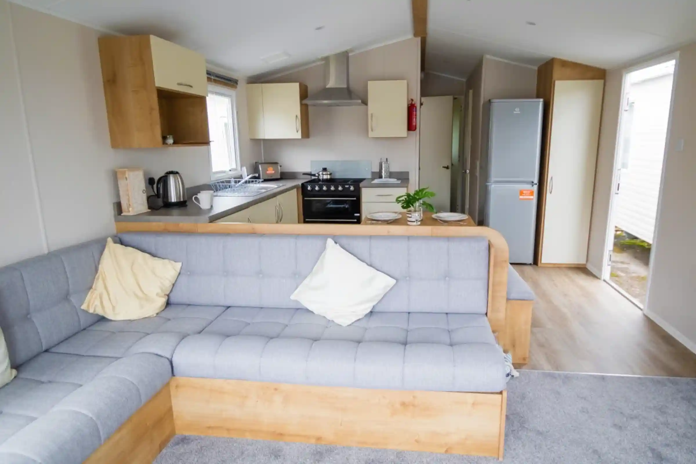 Modern interior of a cozy mobile home featuring a gray sectional sofa with decorative pillows, a compact kitchen with a gas stove and refrigerator, and light wood cabinetry. Natural light enters through the windows, creating a welcoming atmosphere.
