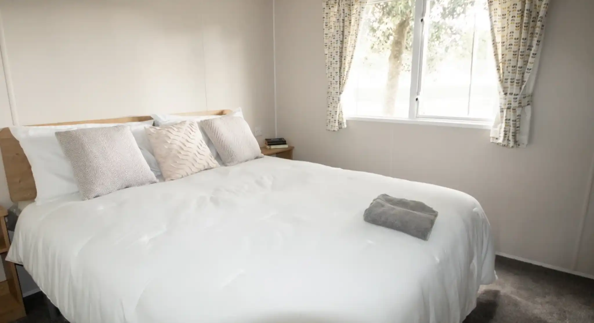 A cozy bedroom featuring a large, neatly made bed with decorative pillows. Natural light filters through a window adorned with patterned curtains, illuminating the neutral-colored walls and carpet. A small bedside table with a drawer is visible next to the bed.