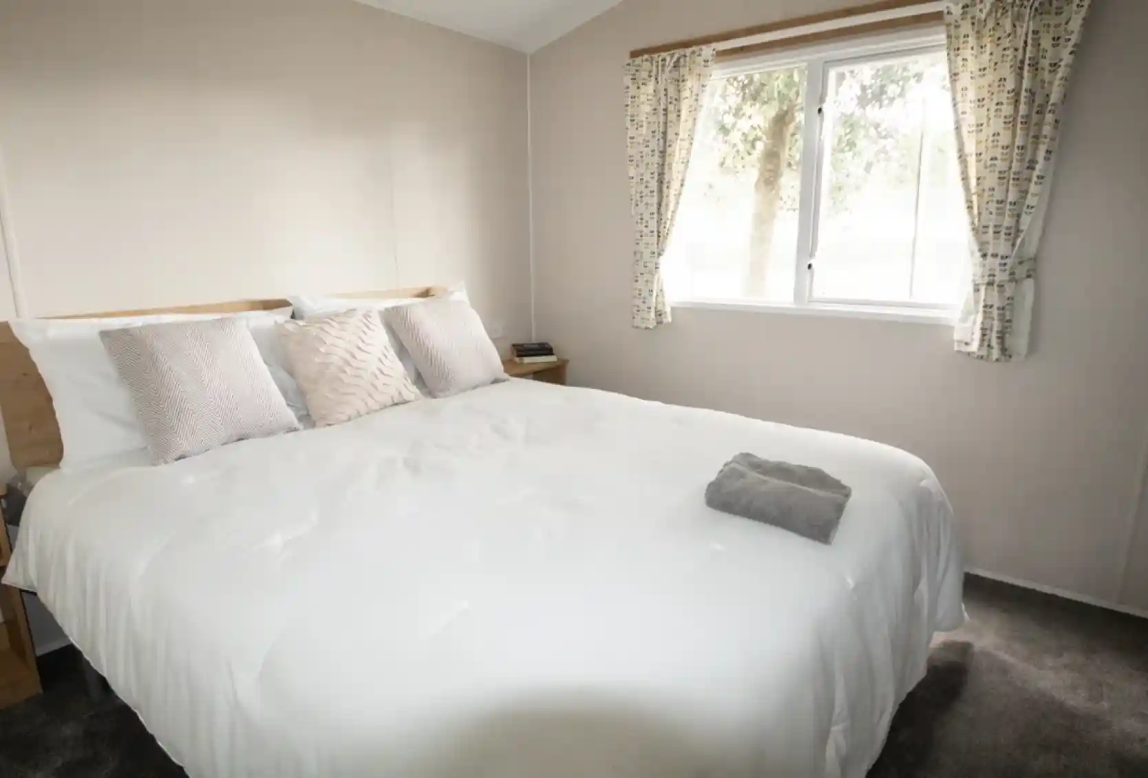 A cozy bedroom featuring a large, neatly made bed with decorative pillows. Natural light filters through a window adorned with patterned curtains, illuminating the neutral-colored walls and carpet. A small bedside table with a drawer is visible next to the bed.