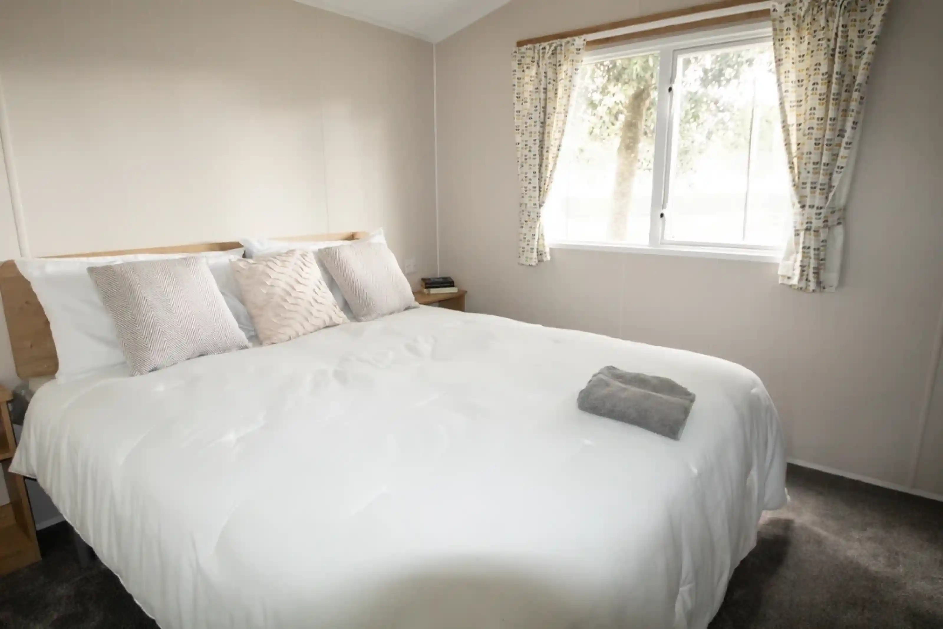 A cozy bedroom featuring a large, neatly made bed with decorative pillows. Natural light filters through a window adorned with patterned curtains, illuminating the neutral-colored walls and carpet. A small bedside table with a drawer is visible next to the bed.