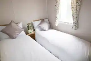 Two twin beds with white linens and decorative pillows are arranged in a small room. A window with patterned curtains offers natural light, and a wooden bedside table holds a stack of green boxes.