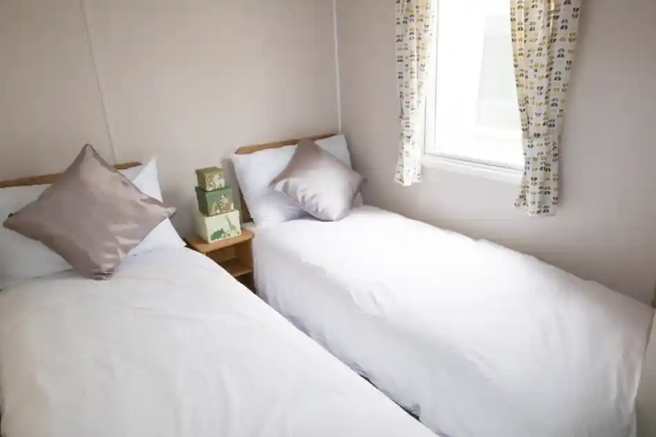 Two twin beds with white linens and decorative pillows are arranged in a small room. A window with patterned curtains offers natural light, and a wooden bedside table holds a stack of green boxes.