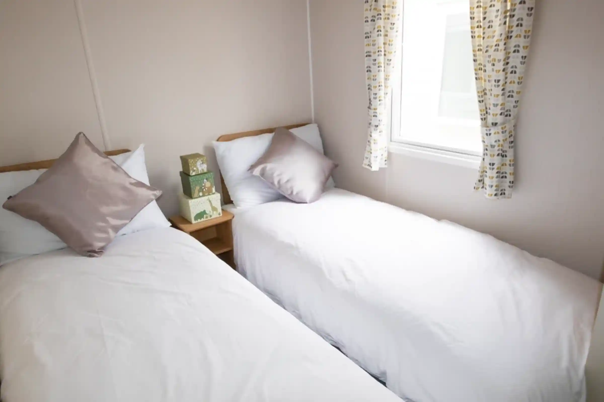 Two twin beds with white linens and decorative pillows are arranged in a small room. A window with patterned curtains offers natural light, and a wooden bedside table holds a stack of green boxes.