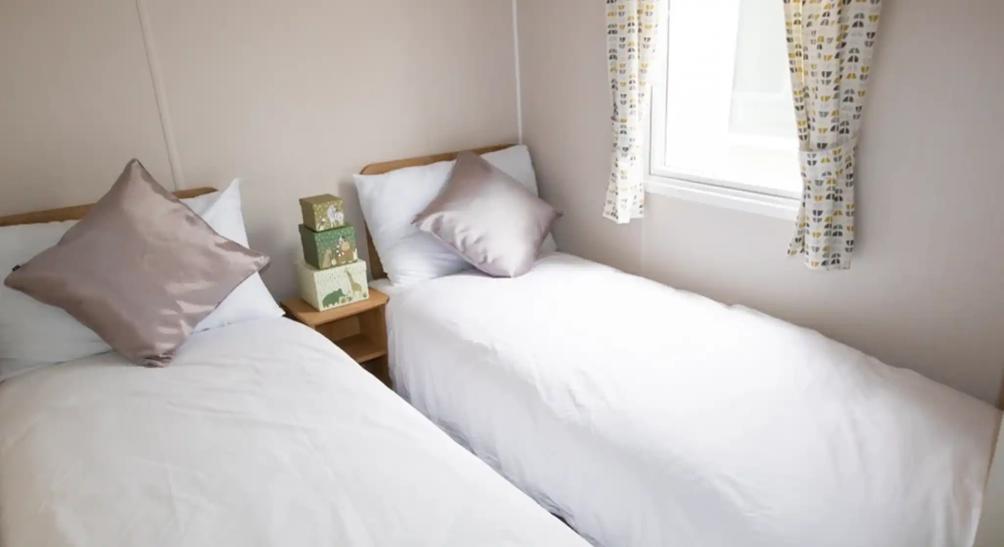 Two twin beds with white linens and decorative pillows are arranged in a small room. A window with patterned curtains offers natural light, and a wooden bedside table holds a stack of green boxes.