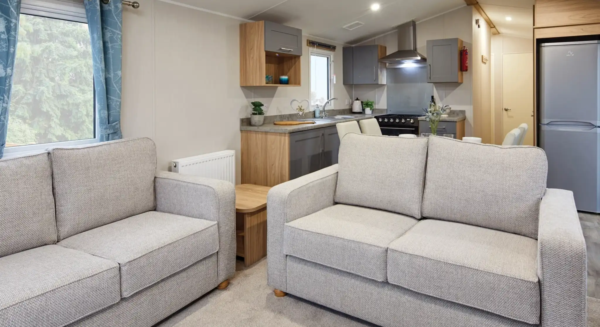 Two grey sofas in a modern living room and kitchen in the background with free-standing fridge freezer, grey cabinetry, stove and sink.