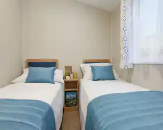 Twin bedroom in a holiday caravan with two single beds, white linens, blue accents, light wood furniture, and a bright, airy feel.