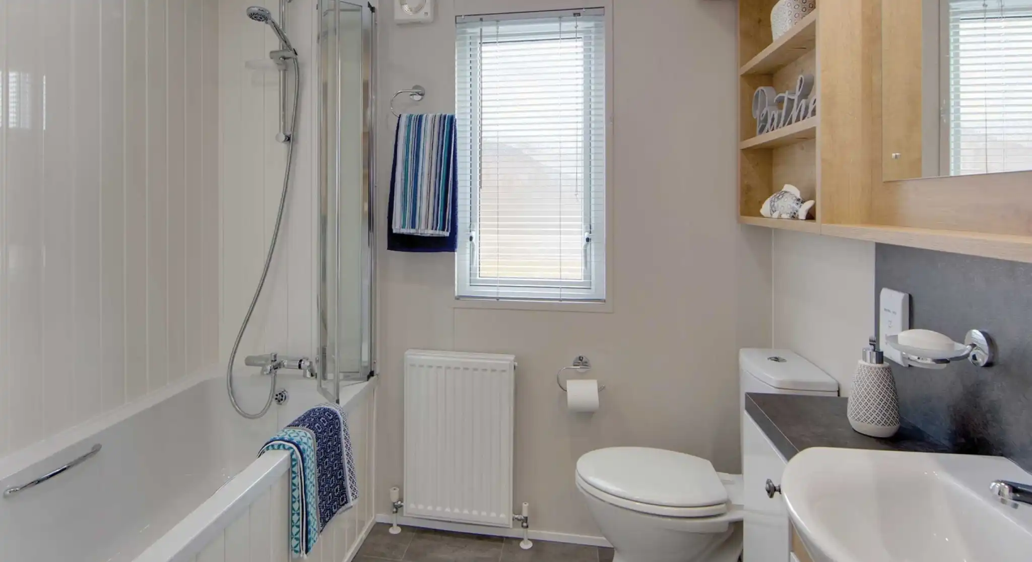 Modern bathroom with gray tile floor, white walls, and a bathtub with glass shower screen. Towels hang near a window above a radiator.