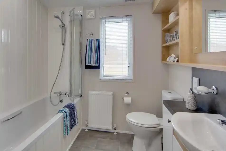 Modern bathroom with gray tile floor, white walls, and a bathtub with glass shower screen. Towels hang near a window above a radiator.