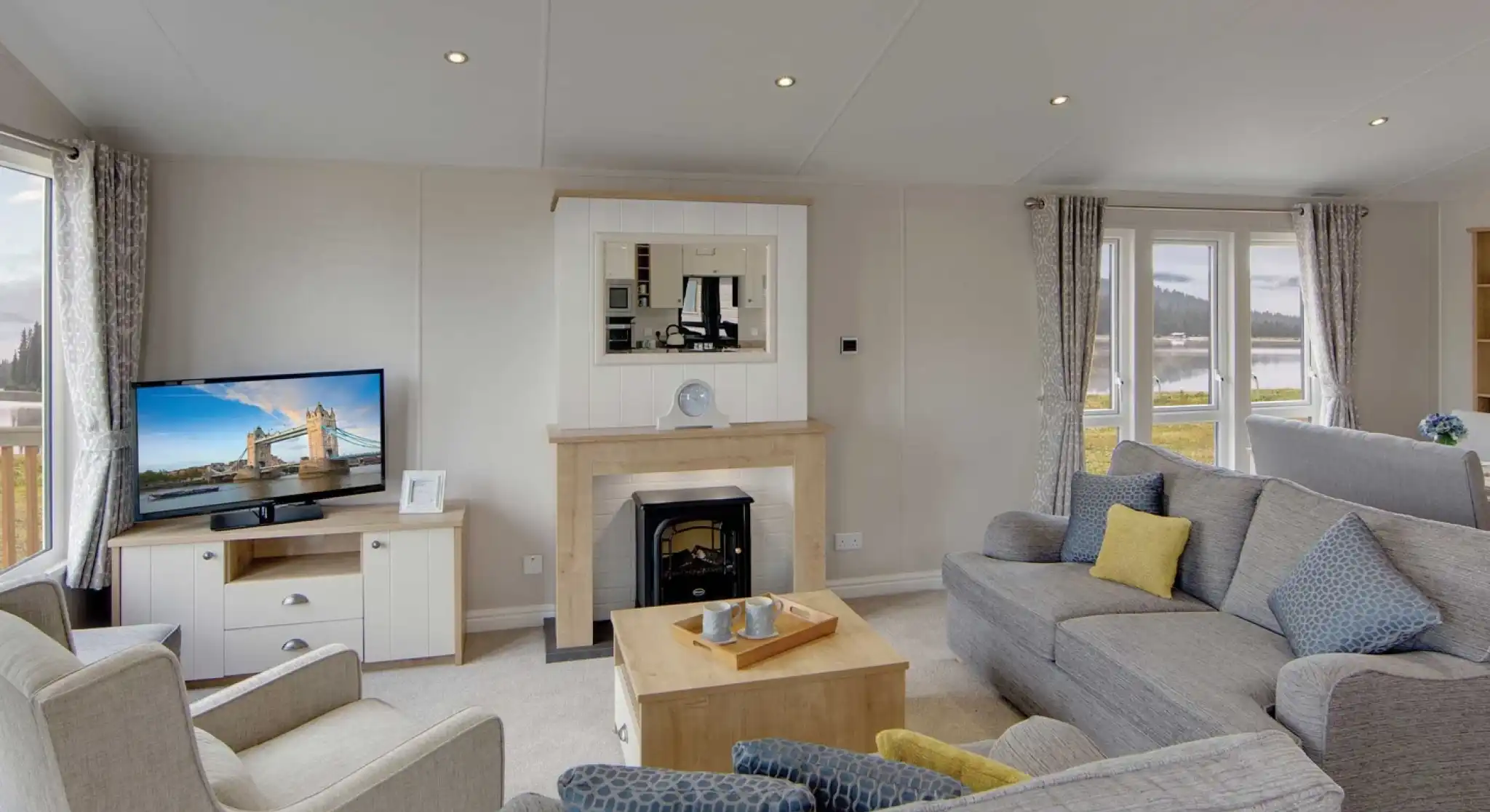 A cozy living room with gray sofas, a beige armchair, and a wooden coffee table. A lit TV displays a cityscape. Large windows show a scenic view.