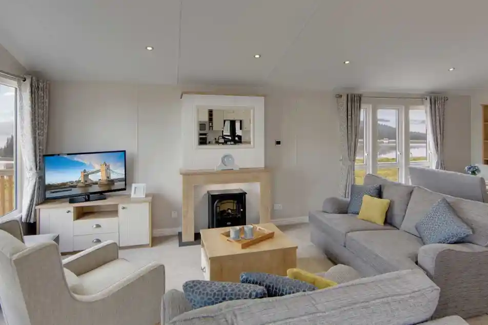 A cozy living room with gray sofas, a beige armchair, and a wooden coffee table. A lit TV displays a cityscape. Large windows show a scenic view.