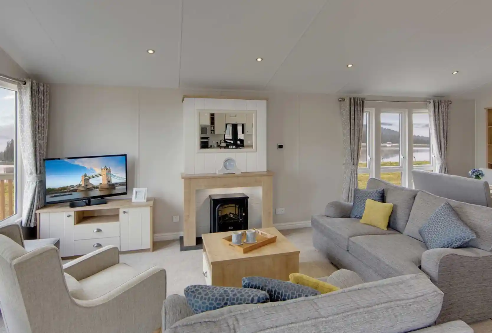 A cozy living room with gray sofas, a beige armchair, and a wooden coffee table. A lit TV displays a cityscape. Large windows show a scenic view.