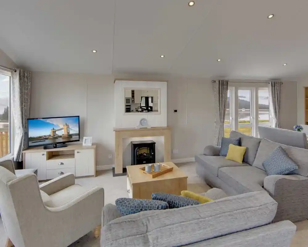 A cozy living room with gray sofas, a beige armchair, and a wooden coffee table. A lit TV displays a cityscape. Large windows show a scenic view.