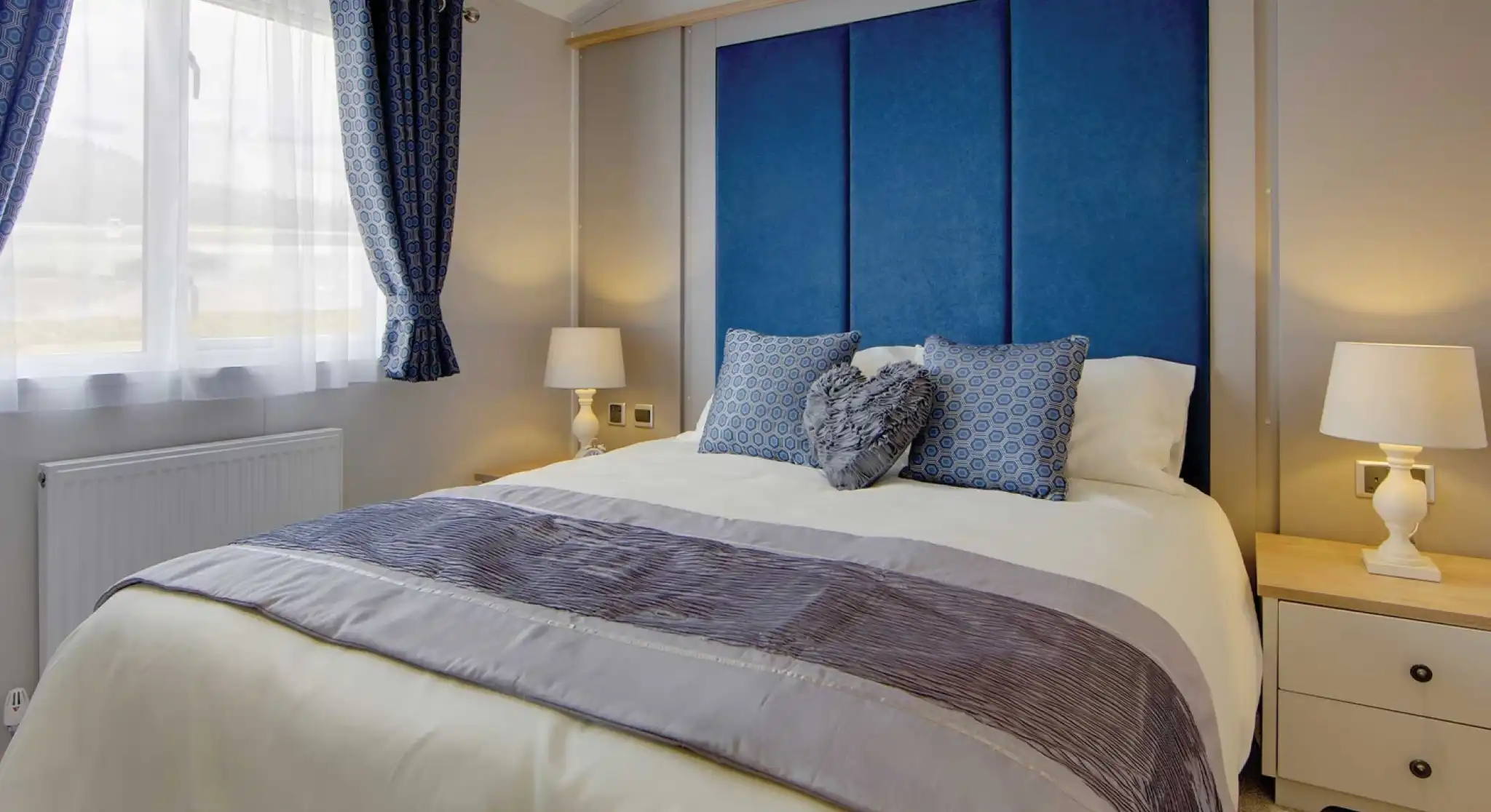 A cozy bedroom with a large bed featuring blue and gray pillows, a blue padded headboard, and light wooden bedside tables with white lamps. Soft natural light filters through blue-patterned curtains, creating a calm and inviting atmosphere.