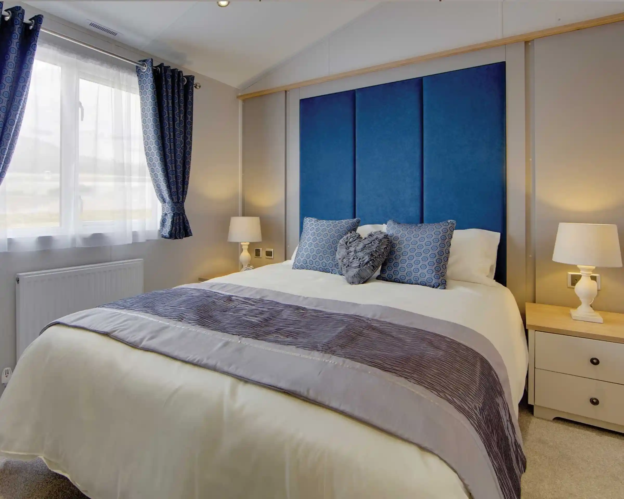 A cozy bedroom with a large bed featuring blue and gray pillows, a blue padded headboard, and light wooden bedside tables with white lamps. Soft natural light filters through blue-patterned curtains, creating a calm and inviting atmosphere.