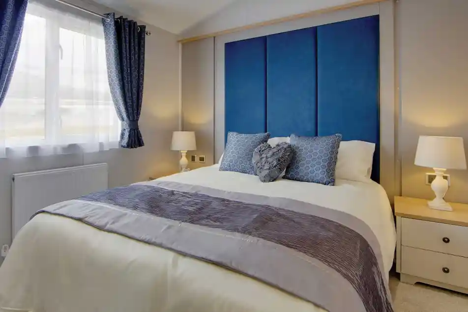 A cozy bedroom with a large bed featuring blue and gray pillows, a blue padded headboard, and light wooden bedside tables with white lamps. Soft natural light filters through blue-patterned curtains, creating a calm and inviting atmosphere.