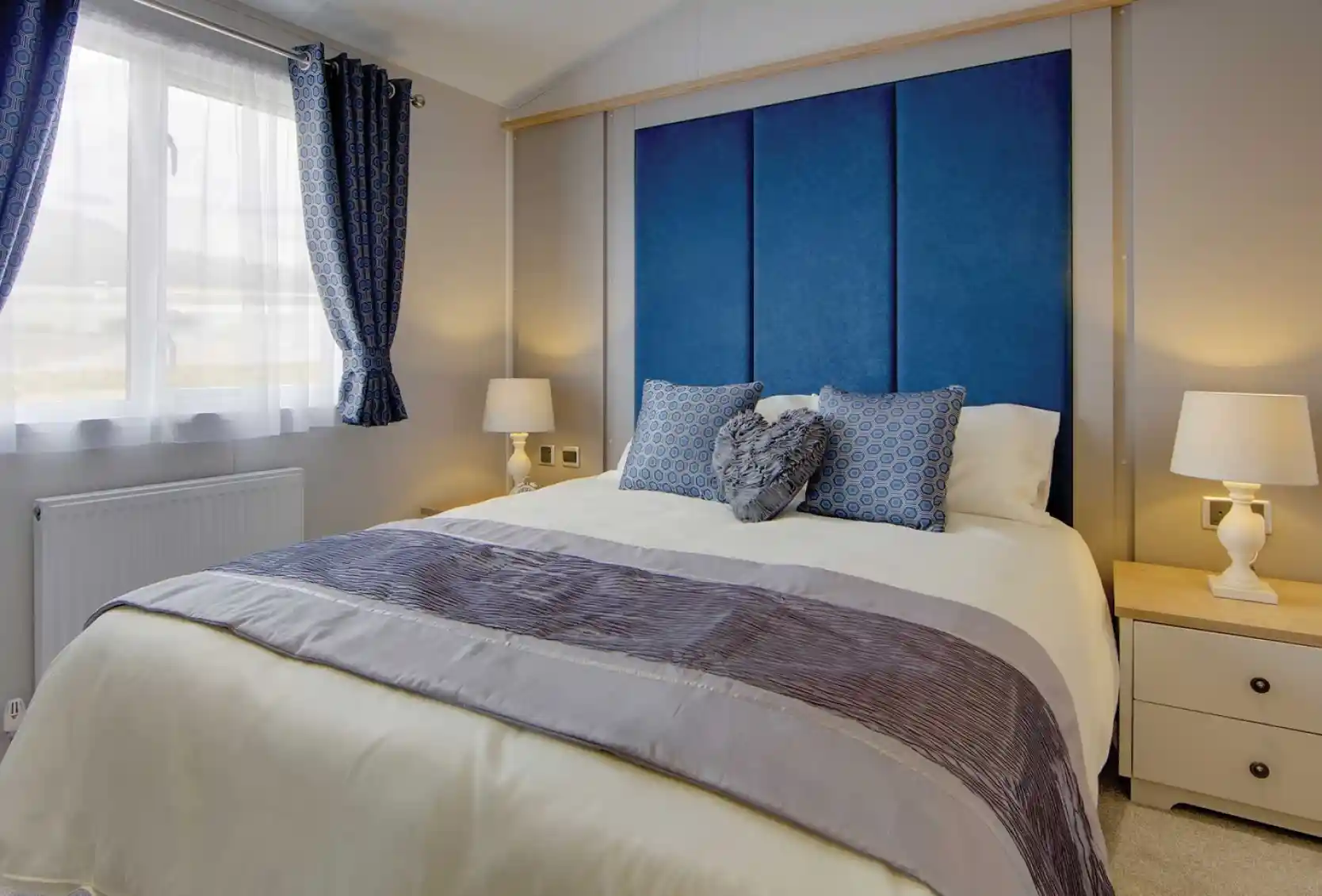 A cozy bedroom with a large bed featuring blue and gray pillows, a blue padded headboard, and light wooden bedside tables with white lamps. Soft natural light filters through blue-patterned curtains, creating a calm and inviting atmosphere.