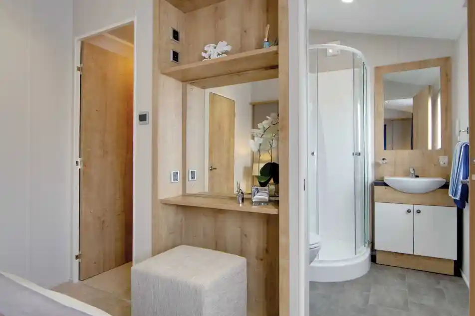 Modern bathroom and dressing area with wood accents. Includes a glass shower, counter with sink, mirror, and upholstered stool, exuding a serene ambiance.