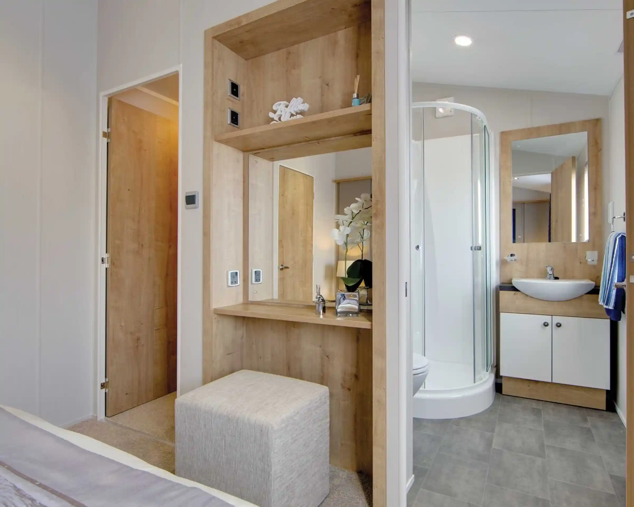 Modern bathroom and dressing area with wood accents. Includes a glass shower, counter with sink, mirror, and upholstered stool, exuding a serene ambiance.