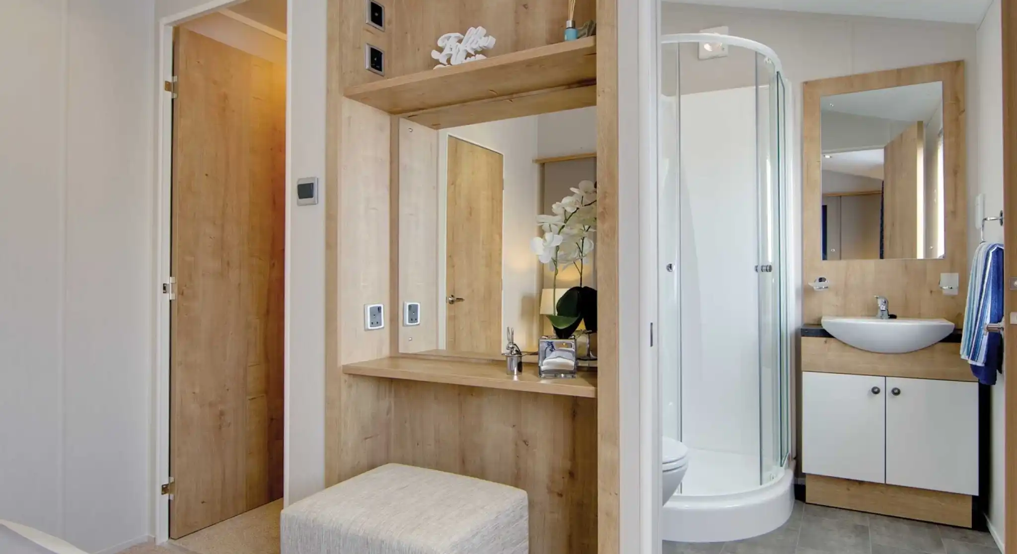 Modern bathroom and dressing area with wood accents. Includes a glass shower, counter with sink, mirror, and upholstered stool, exuding a serene ambiance.