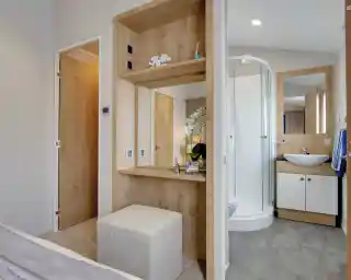 Modern bathroom and dressing area with wood accents. Includes a glass shower, counter with sink, mirror, and upholstered stool, exuding a serene ambiance.