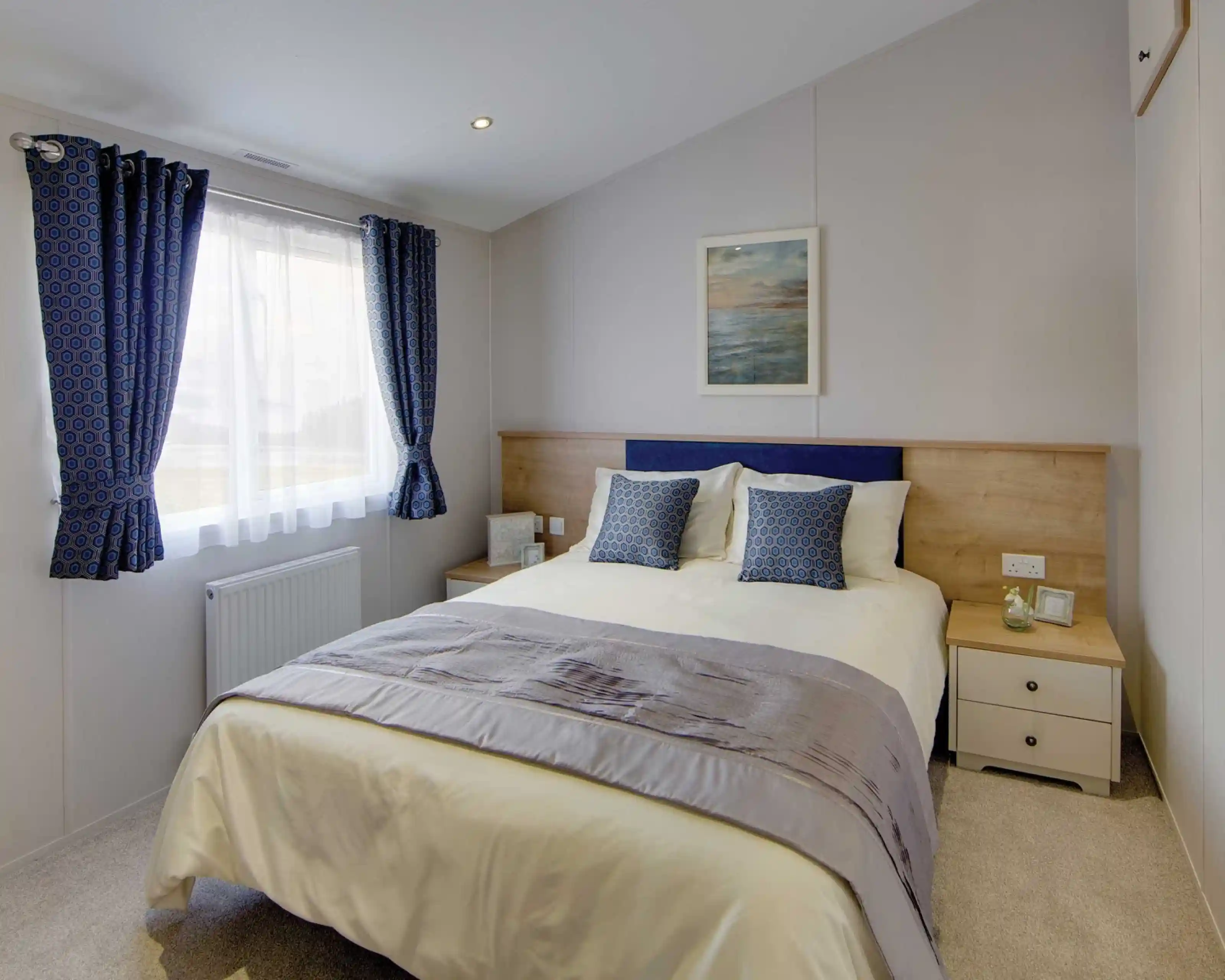 Bright bedroom with a double bed, two blue patterned cushions, wooden headboard, and ocean-themed wall art. Blue curtains frame a window. Cozy ambiance.