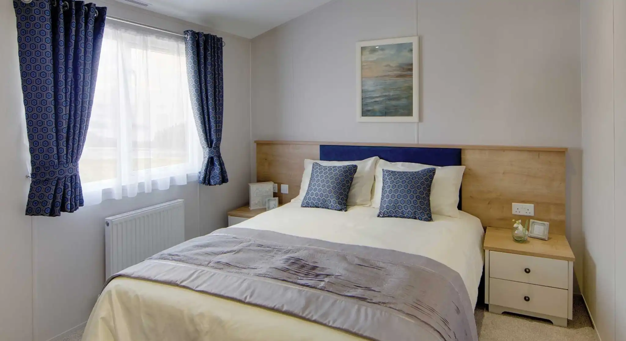 Bright bedroom with a double bed, two blue patterned cushions, wooden headboard, and ocean-themed wall art. Blue curtains frame a window. Cozy ambiance.