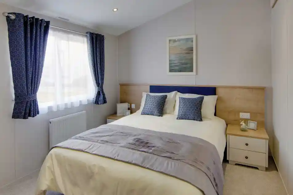 Bright bedroom with a double bed, two blue patterned cushions, wooden headboard, and ocean-themed wall art. Blue curtains frame a window. Cozy ambiance.