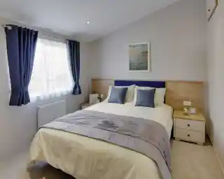 Bright bedroom with a double bed, two blue patterned cushions, wooden headboard, and ocean-themed wall art. Blue curtains frame a window. Cozy ambiance.