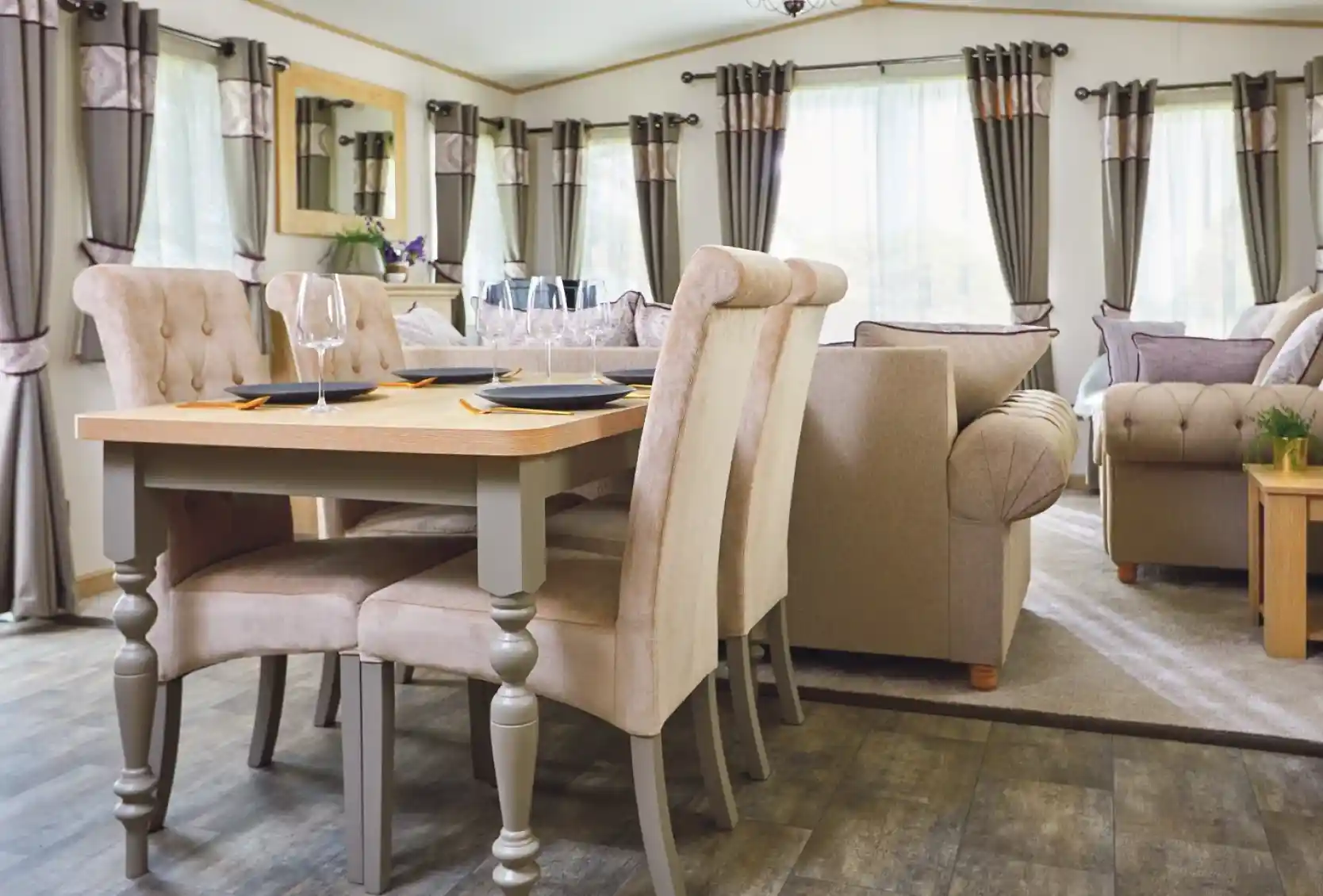 Modern dining area featuring a wooden table with four beige upholstered chairs. A cozy living space in the background includes a sofa, large windows with elegant curtains, and soft lighting.