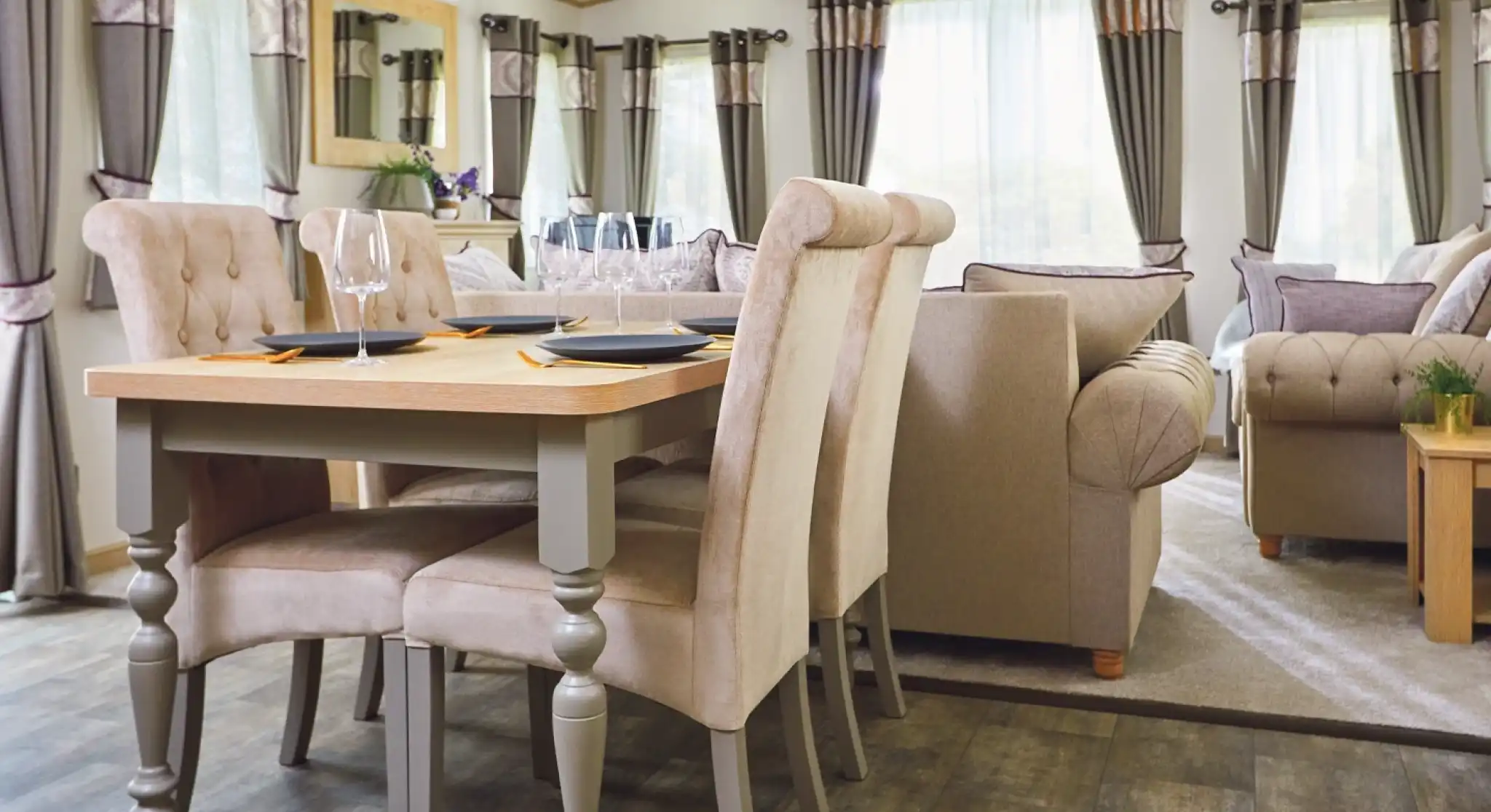 Modern dining area featuring a wooden table with four beige upholstered chairs. A cozy living space in the background includes a sofa, large windows with elegant curtains, and soft lighting.
