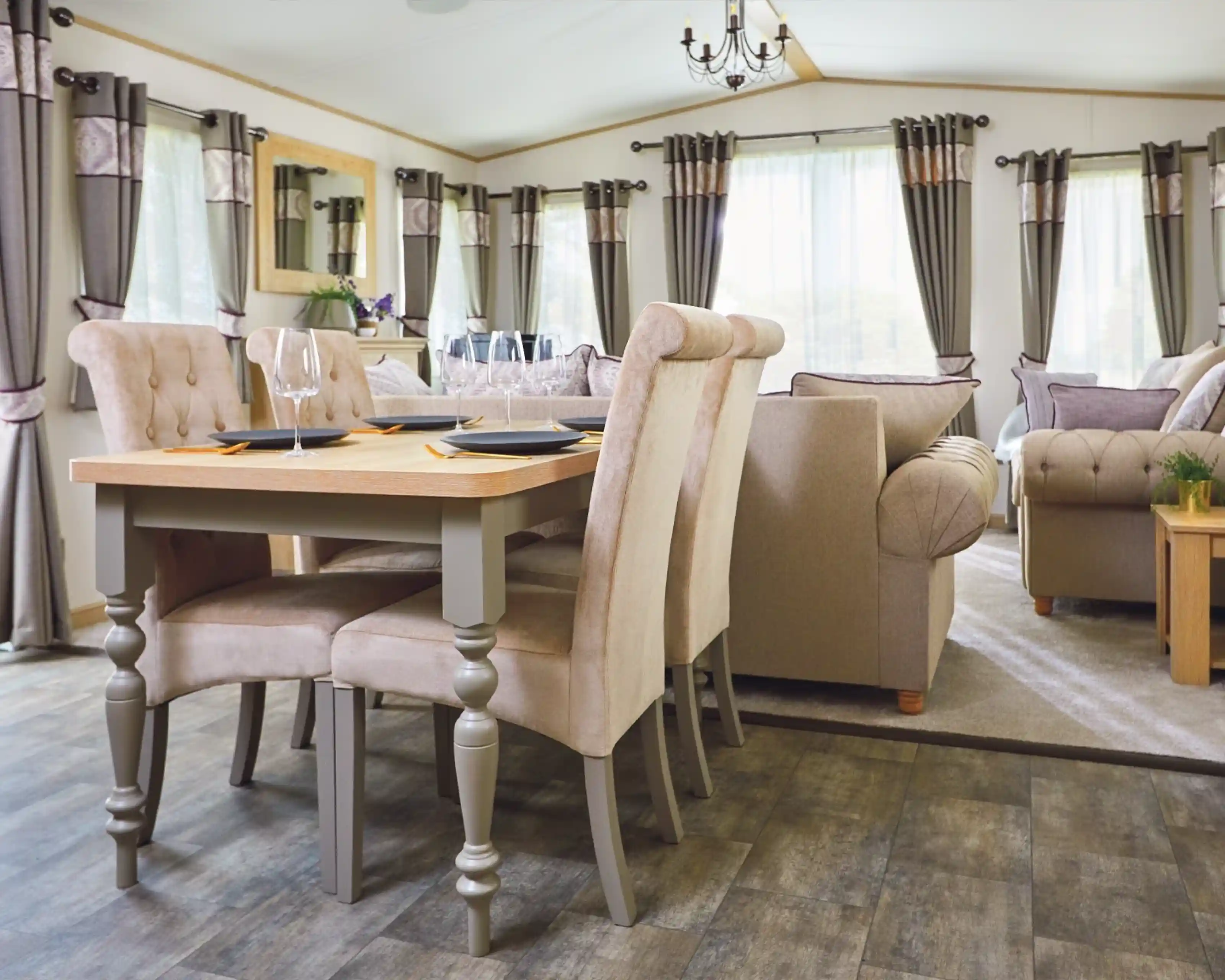 Modern dining area featuring a wooden table with four beige upholstered chairs. A cozy living space in the background includes a sofa, large windows with elegant curtains, and soft lighting.