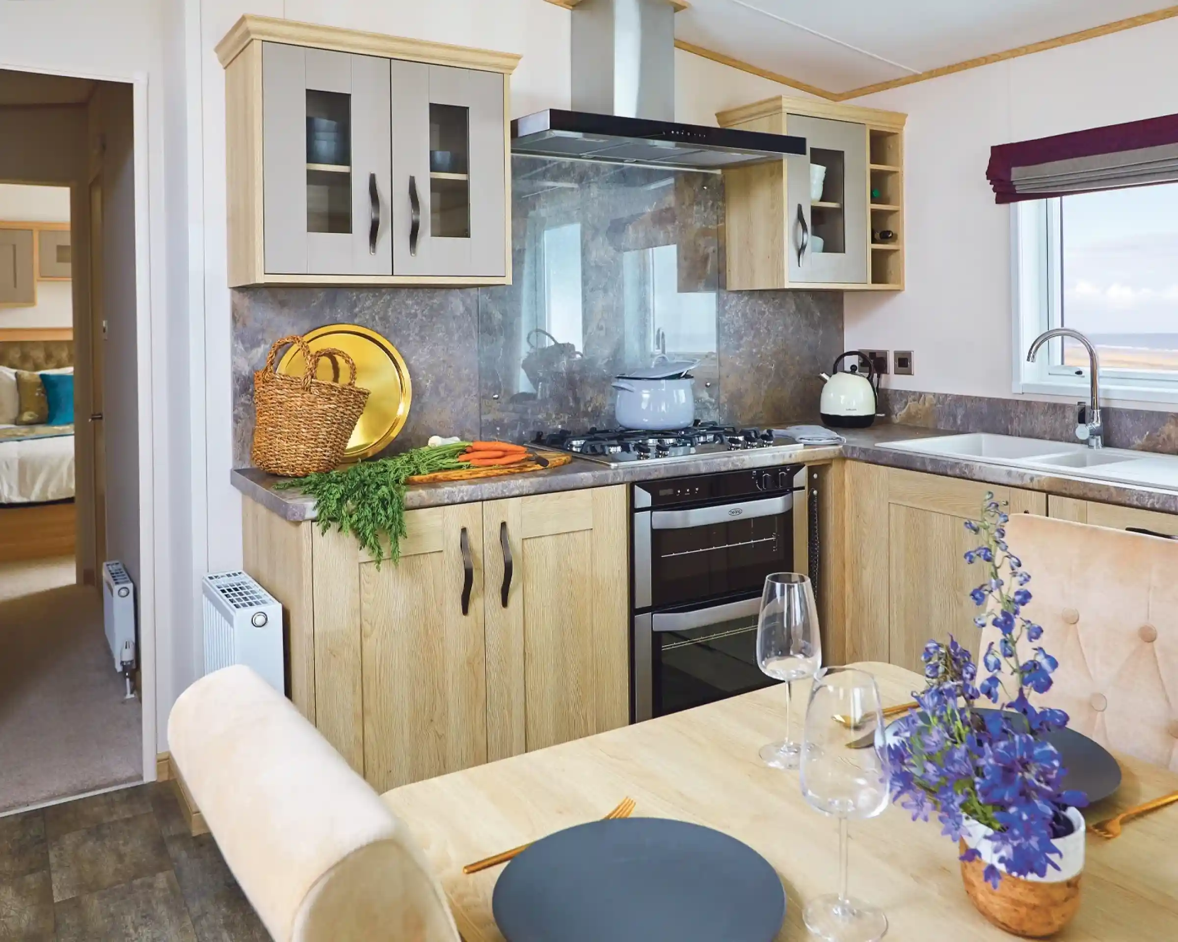 A modern kitchen with light wood cabinetry, a gas stove, and a countertop. A dining table is set with plates and wine glasses, while a bouquet of purple flowers adds a decorative touch. Fresh vegetables are placed on the counter, and a view of the outdoors is visible through a window.