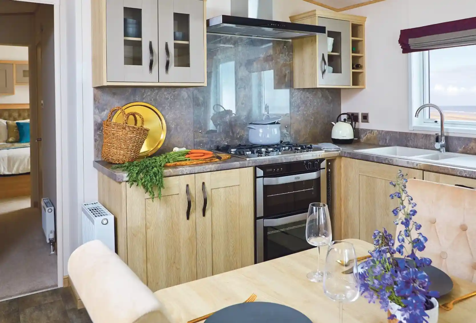 A modern kitchen with light wood cabinetry, a gas stove, and a countertop. A dining table is set with plates and wine glasses, while a bouquet of purple flowers adds a decorative touch. Fresh vegetables are placed on the counter, and a view of the outdoors is visible through a window.